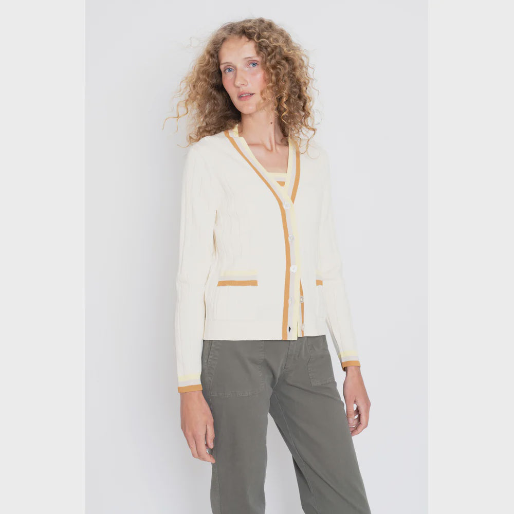 Someone with curly hair models The Dede Cream/Hazelnut by Kule, featuring a cream cotton cardigan with yellow accents over a matching top and olive green pants. Standing against a white background, this lightweight piece adds effortless charm for spring/summer layering.