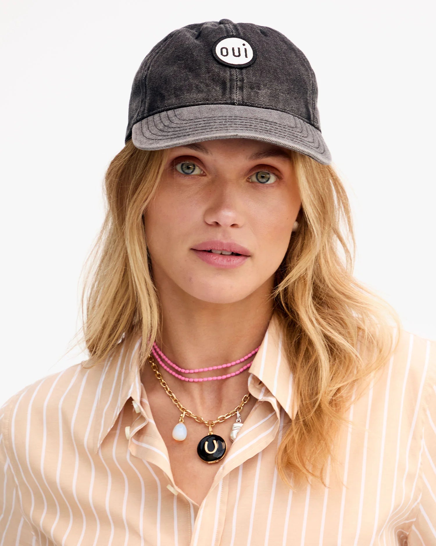 A woman with long blonde hair wears the Clare Vivier Denim Baseball Hat, featuring an adjustable closure and "oui" embroidery, styled with a beige and white striped shirt and layered necklaces. One size fits most.