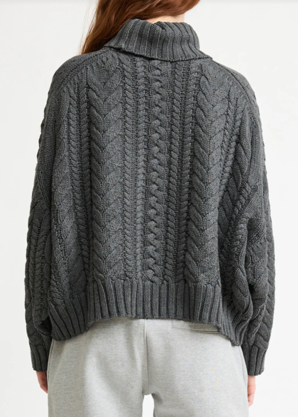 A person is seen from the back wearing the Denimist CABLE TURTLENECK SWEATER in chunky, dark gray cable knit, paired with light gray sweatpants.
