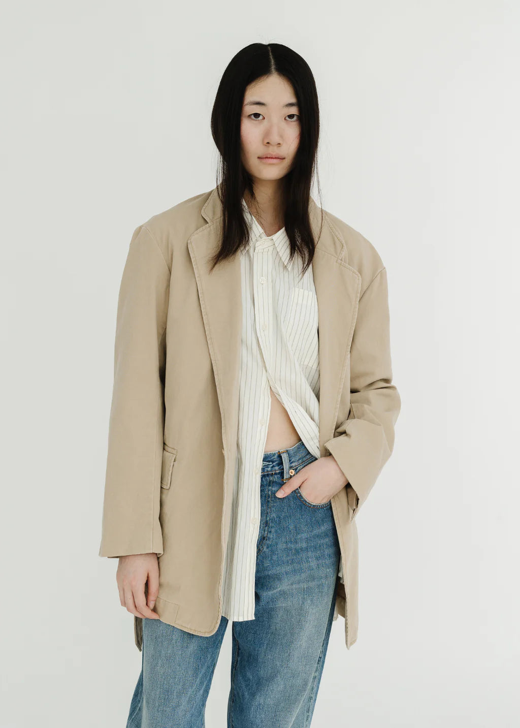 A person with long black hair stands against a plain white background, wearing Denimist's DECONSTRUCTED BLAZER WITH BELT in beige, styled over a white striped shirt and blue jeans.