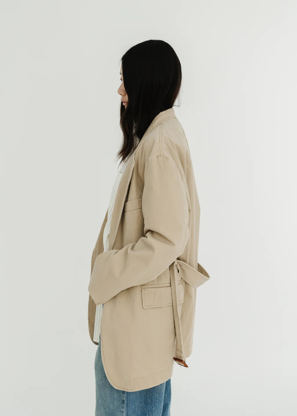 A woman with long dark hair stands in profile against a plain white background, wearing the Denimist DECONSTRUCTED BLAZER WITH BELT—an oversized beige blazer—paired with blue jeans.