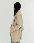 A woman with long dark hair stands in profile against a plain white background, wearing the Denimist DECONSTRUCTED BLAZER WITH BELT—an oversized beige blazer—paired with blue jeans.