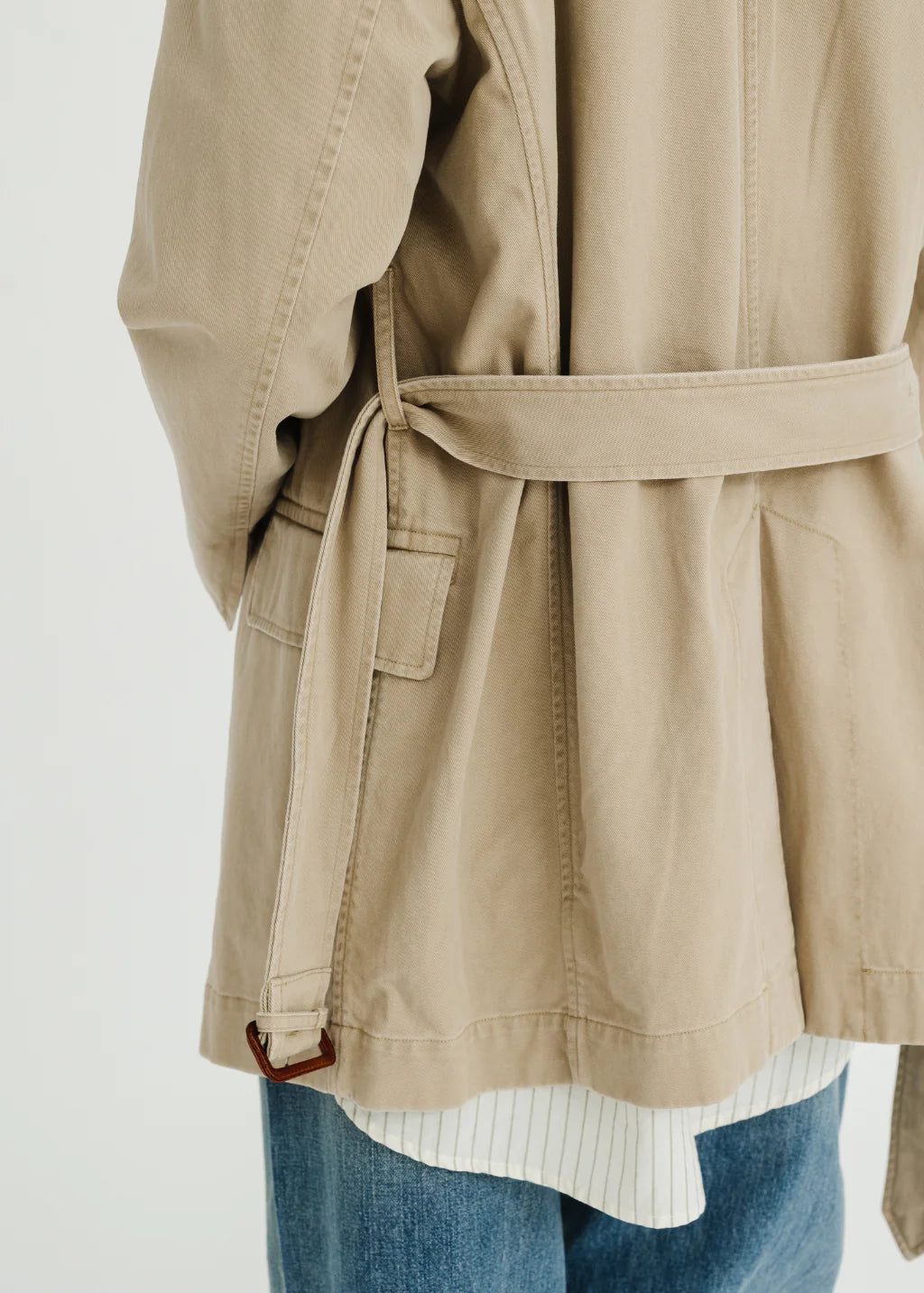 A person is shown from the back against a plain background, wearing the Denimist DECONSTRUCTED BLAZER WITH BELT—an oversized beige piece—layered over a white shirt with thin stripes and blue jeans.