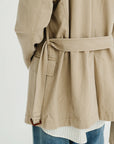 A person is shown from the back against a plain background, wearing the Denimist DECONSTRUCTED BLAZER WITH BELT—an oversized beige piece—layered over a white shirt with thin stripes and blue jeans.