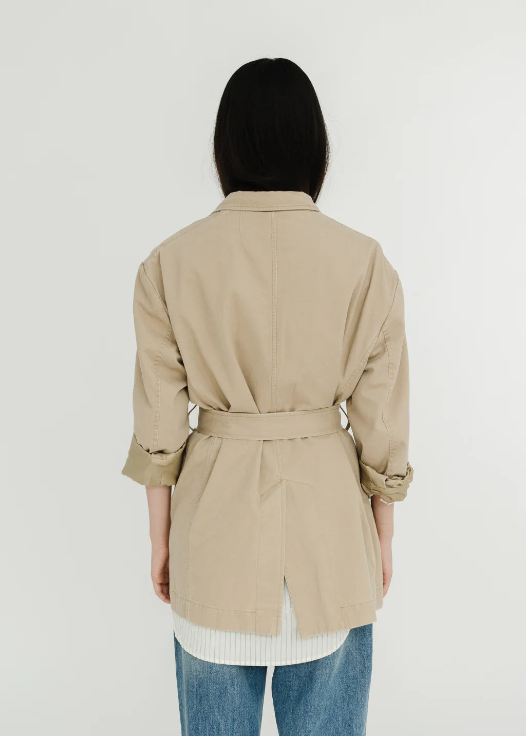A person with long dark hair, seen from behind, is wearing the Denimist DECONSTRUCTED BLAZER WITH BELT in beige over a white shirt and blue jeans, standing against a plain white background.