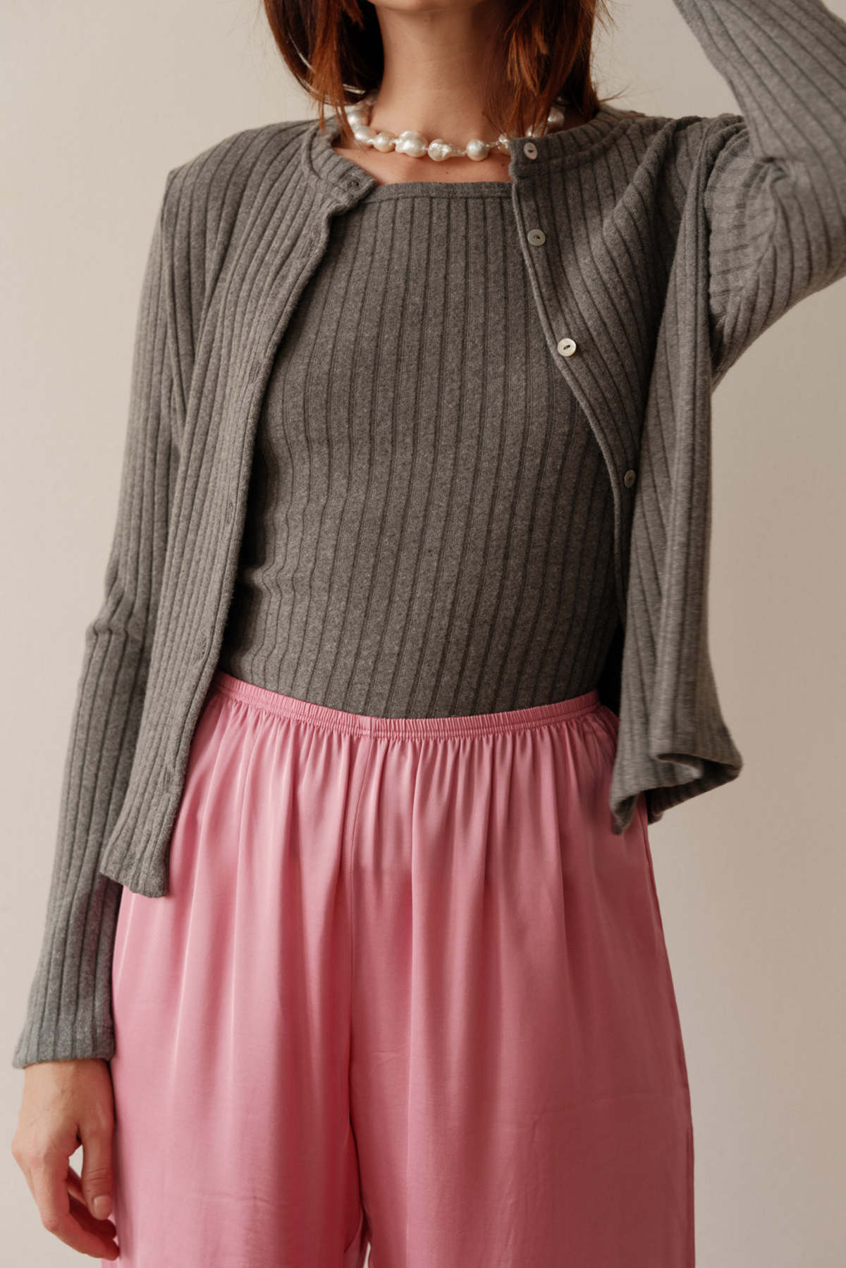 A person wears Donni’s The Sweater Rib Cardi with ribbed texture and mother of pearl buttons, layered over a matching top, styled with loose light pink pants and a chunky pearl necklace. Their face is not visible.