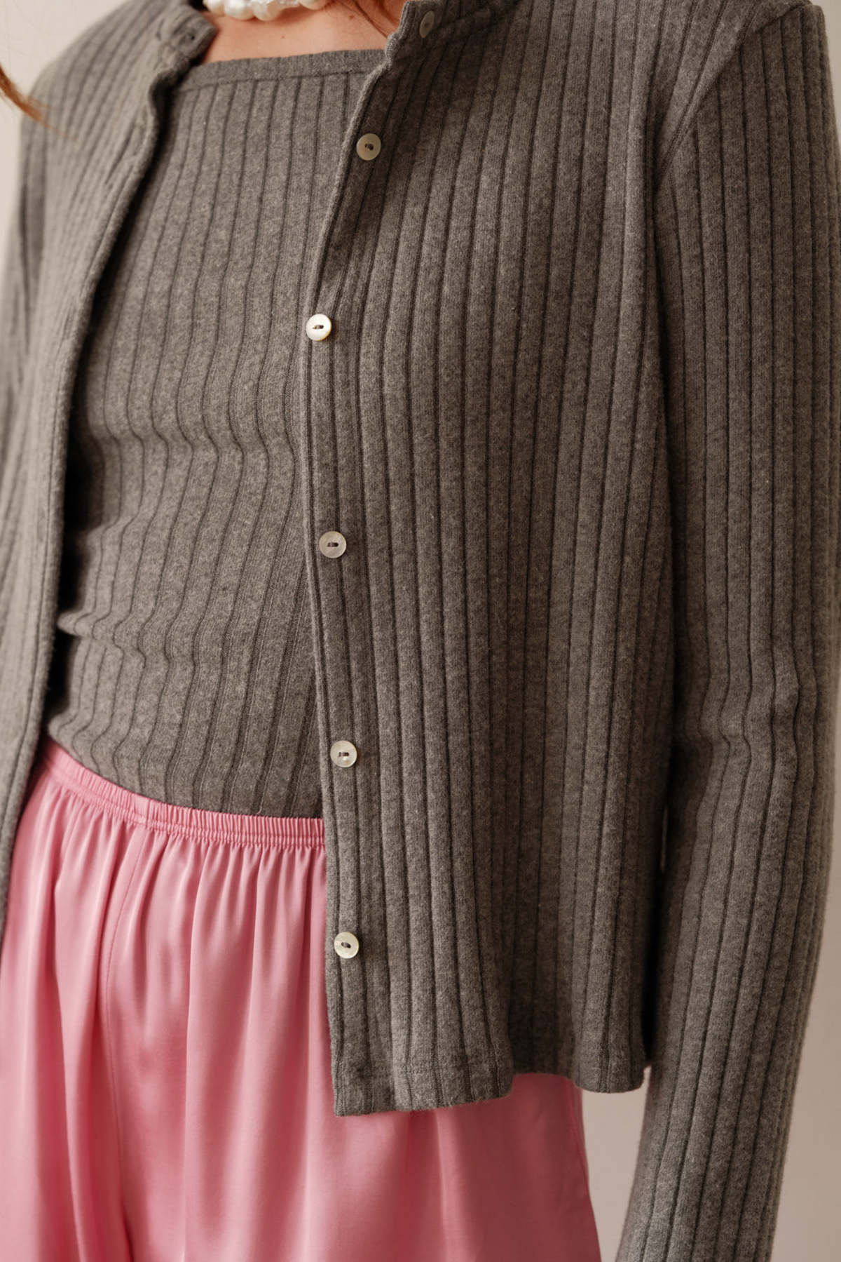 A person wears Donni's The Sweater Rib Cardi in gray with mother of pearl buttons over a matching ribbed top, paired with pink satin pajama pants. Only the torso and neck show, highlighting the cardi’s cozy ribbed texture.