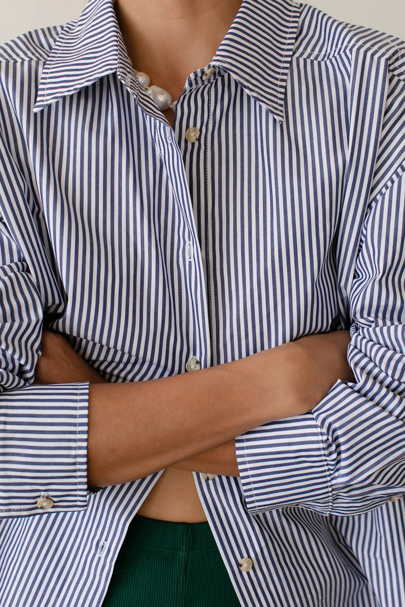 A person with crossed arms wears Donni&#39;s The Stripe Pop Shirt in blue and white cotton poplin stripes, partially unbuttoned to reveal a green garment underneath. The shirt features unique tortoise-like buttons.