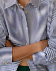 A person with crossed arms wears Donni's The Stripe Pop Shirt in blue and white cotton poplin stripes, partially unbuttoned to reveal a green garment underneath. The shirt features unique tortoise-like buttons.