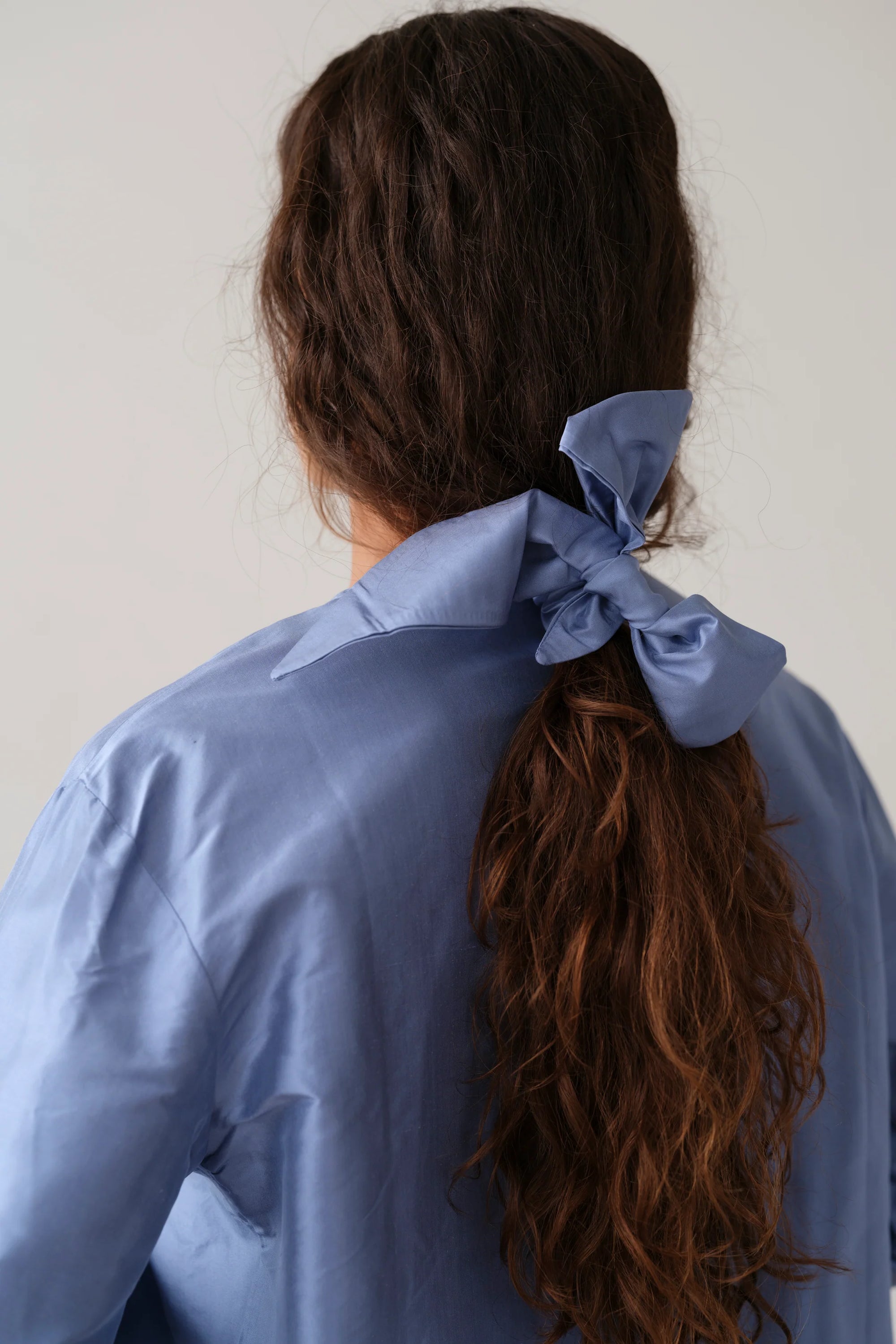 A person with long, wavy brown hair is seen from behind, wearing a light blue shirt and securing their ponytail with Donni's The Taffeta Bow Cornflower Blue accessory.