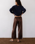 A person stands indoors with hands on hips, wearing Donni's The Silk Stripe Crop Pant—brown, straight-leg, cropped silk pants with white side stripes—paired with a navy sweater and slippers against a plain light wall.