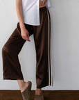 A person stands against a white wall, one hand on their hip, wearing Donni's Silk Stripe Crop Pant in brown satin with a beige side stripe and drawstring waist, paired with a white short-sleeve button-up shirt and brown slippers.