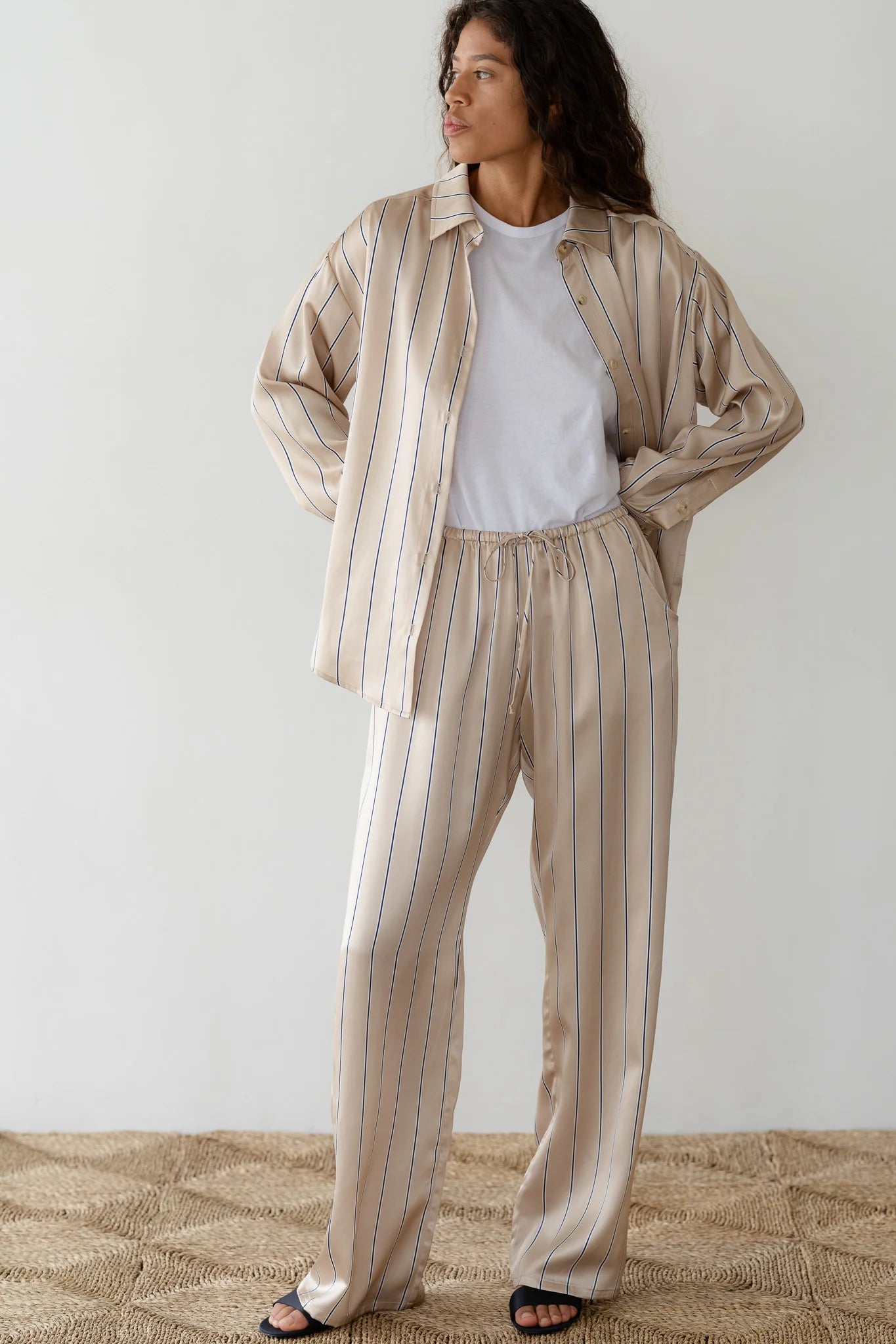 In a minimal setting, a person with long, curly hair wears The Donni Silk Pinstripe Drawstring Pant in beige over a white T-shirt. They gaze to the side with hands in pockets, standing on a textured rug, exuding relaxation against a plain wall background.