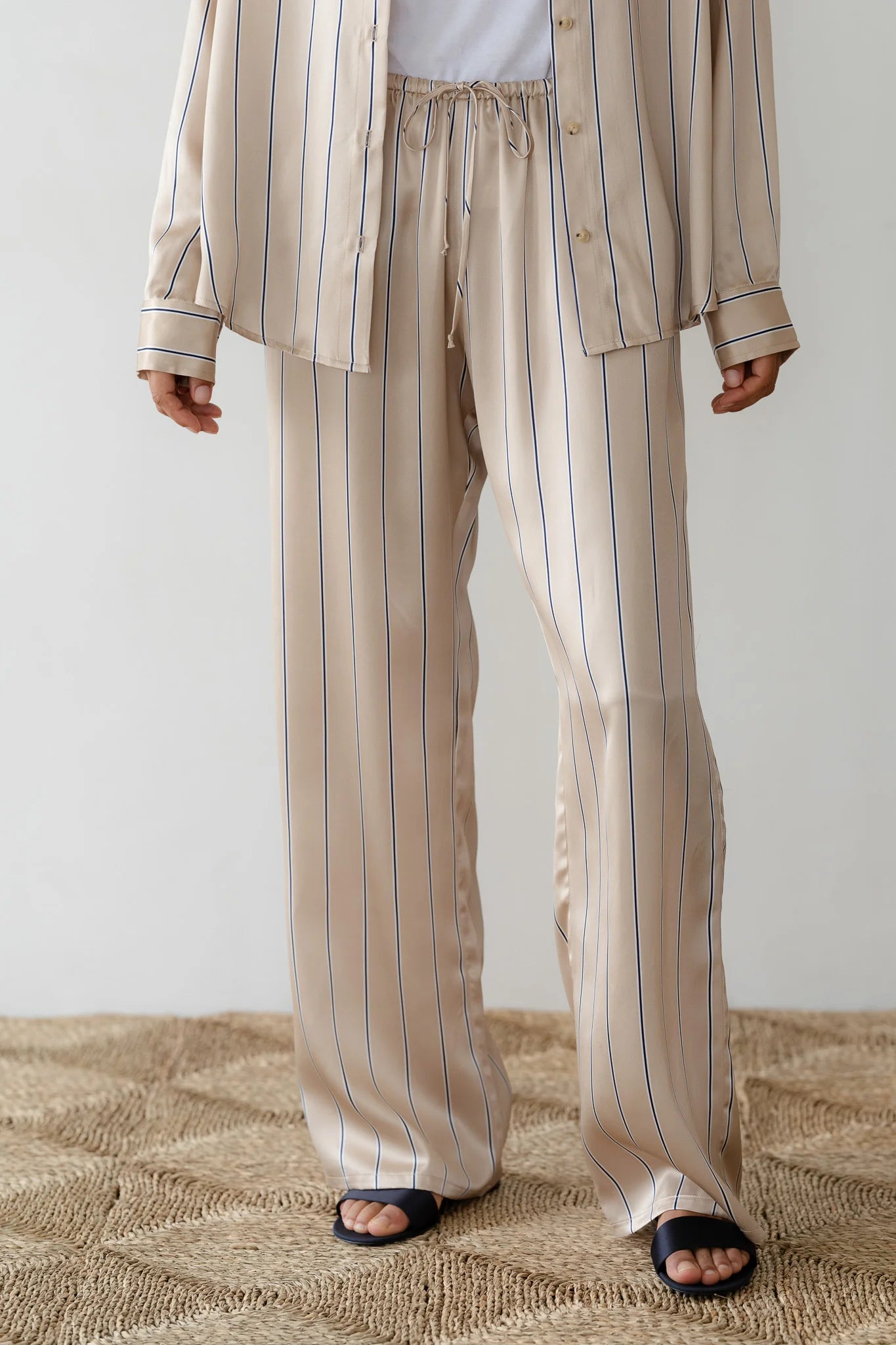 A person, dressed in Donni's Silk Pinstripe Drawstring Pant resembling elegance, wears beige satin pajamas with blue vertical stripes and a relaxed fit. The button-up shirt pairs effortlessly with loose-fitting pants above black open-toe slippers on a textured beige rug.