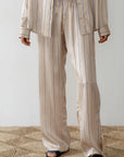 A person, dressed in Donni's Silk Pinstripe Drawstring Pant resembling elegance, wears beige satin pajamas with blue vertical stripes and a relaxed fit. The button-up shirt pairs effortlessly with loose-fitting pants above black open-toe slippers on a textured beige rug.