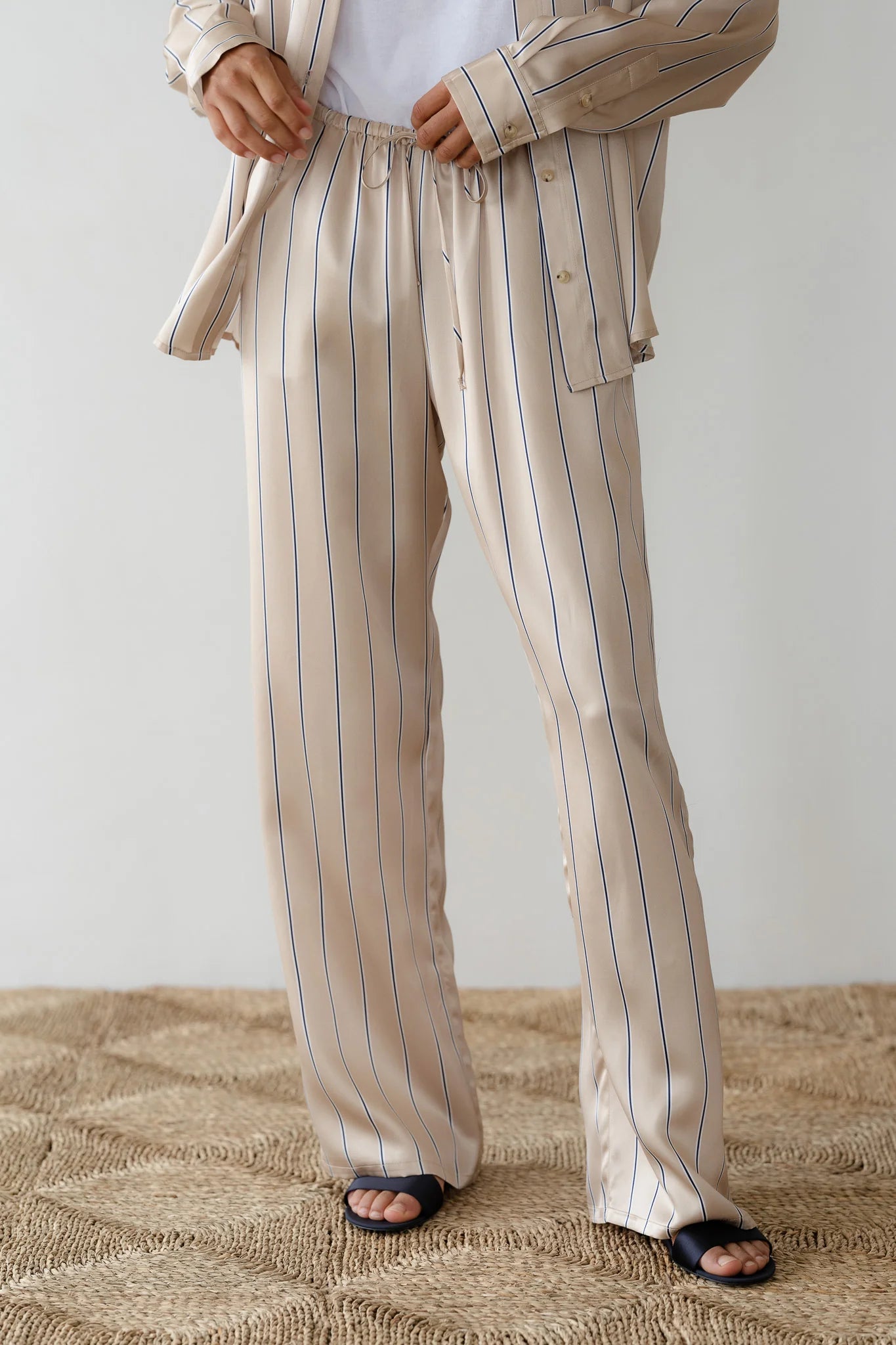 Someone wearing Donni's The Silk Pinstripe Drawstring Pant in relaxed fit, 100% silk beige with vertical black stripes, stands on a textured, beige rug. They pair it with an open matching pajama shirt and black slip-on sandals against a plain light-colored wall.