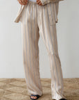 Someone wearing Donni's The Silk Pinstripe Drawstring Pant in relaxed fit, 100% silk beige with vertical black stripes, stands on a textured, beige rug. They pair it with an open matching pajama shirt and black slip-on sandals against a plain light-colored wall.