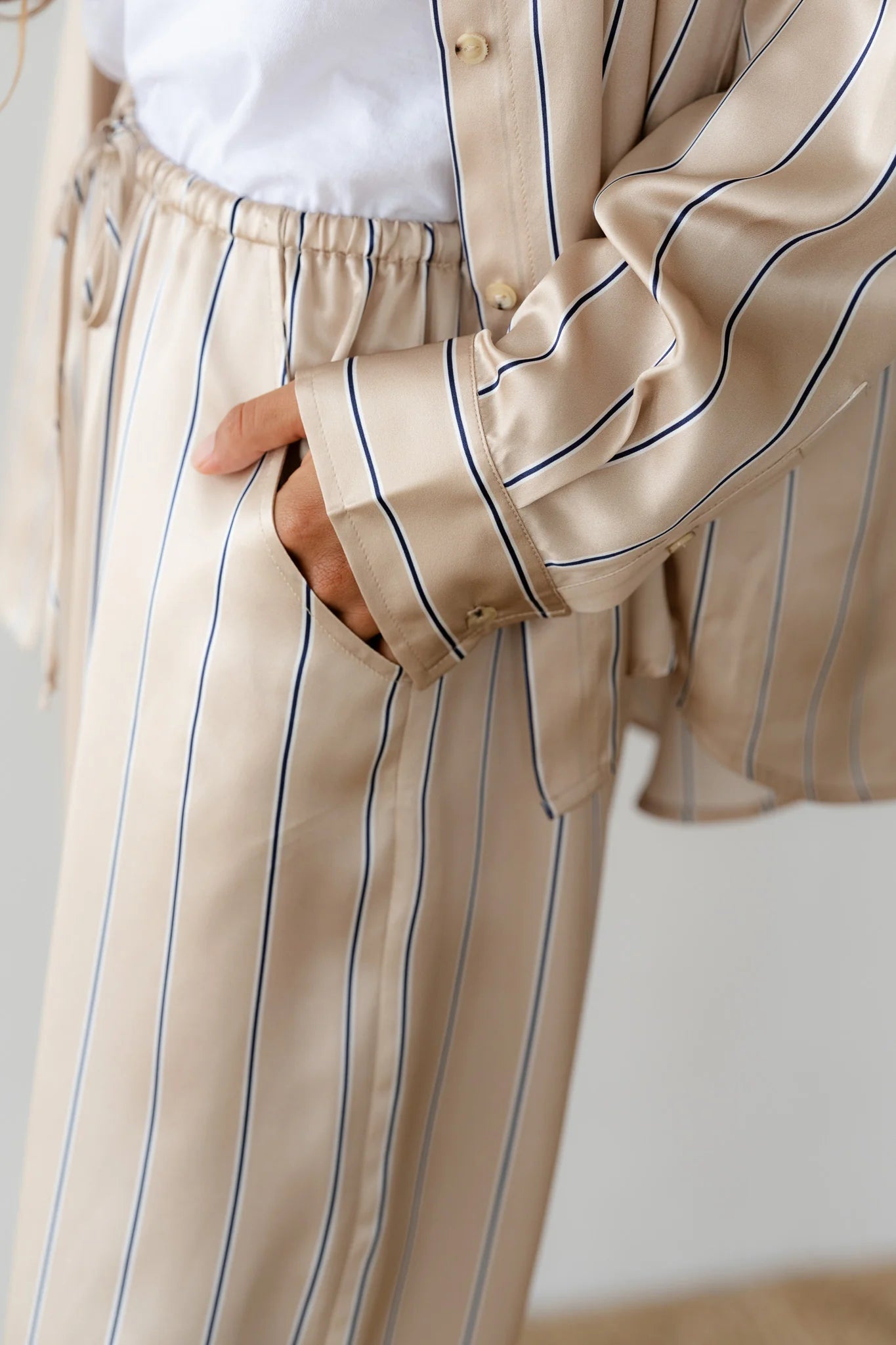 Dressed in relaxed fit beige striped pajamas over a white tee, someone exudes classic style with navy blue pinstripes. The Donni Silk Pinstripe Drawstring Pant adds a touch of luxury as they effortlessly slip one hand into their pocket.