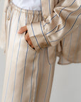 Dressed in relaxed fit beige striped pajamas over a white tee, someone exudes classic style with navy blue pinstripes. The Donni Silk Pinstripe Drawstring Pant adds a touch of luxury as they effortlessly slip one hand into their pocket.