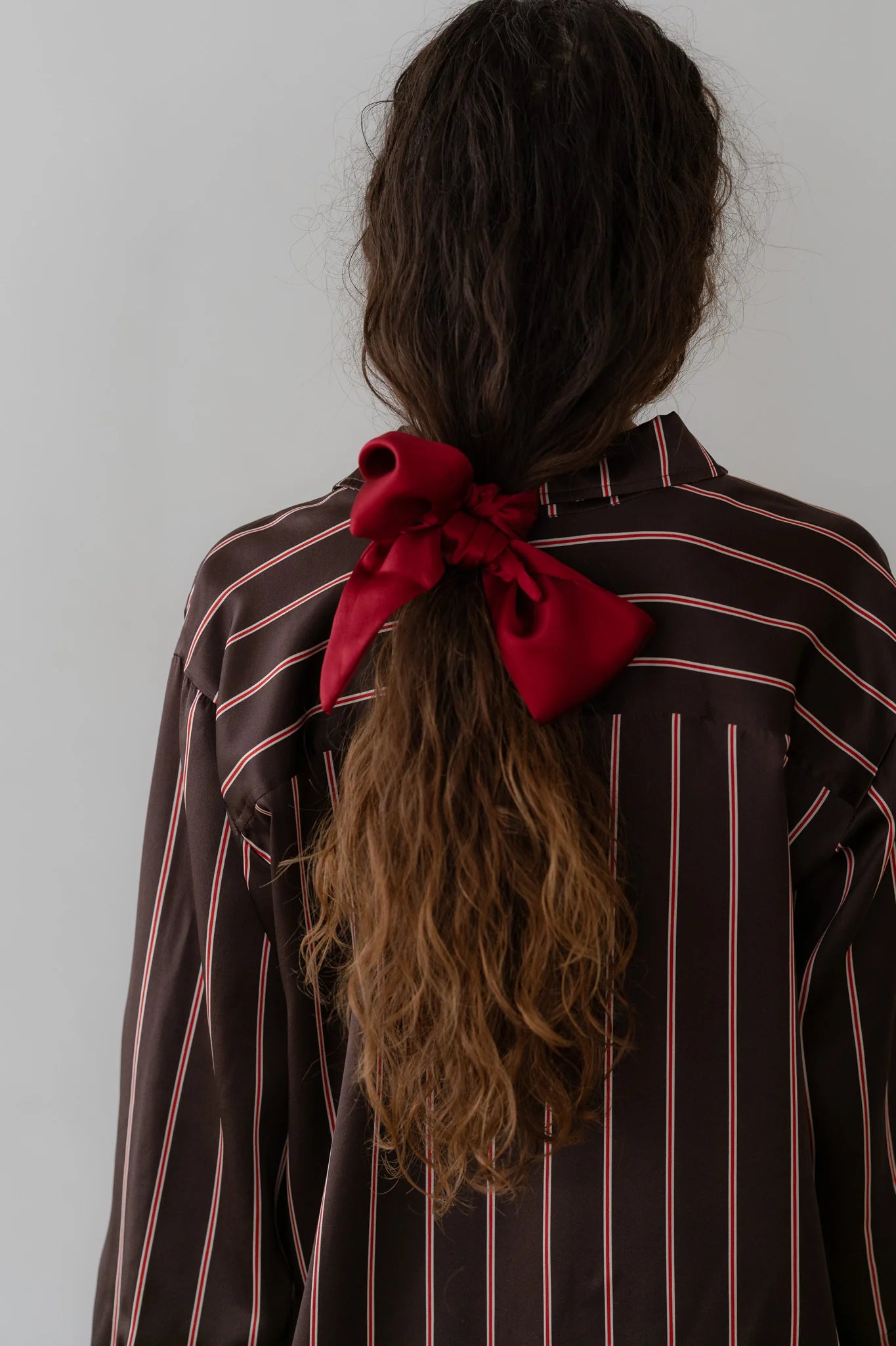 A person with long, wavy hair tied back with The Silk Bow by Donni. They are wearing a dark shirt featuring thin pink and white vertical stripes. The accessory stands out against the plain gray background, accentuating the ensemble's elegance.