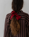 A person with long, wavy hair tied back with The Silk Bow by Donni. They are wearing a dark shirt featuring thin pink and white vertical stripes. The accessory stands out against the plain gray background, accentuating the ensemble's elegance.