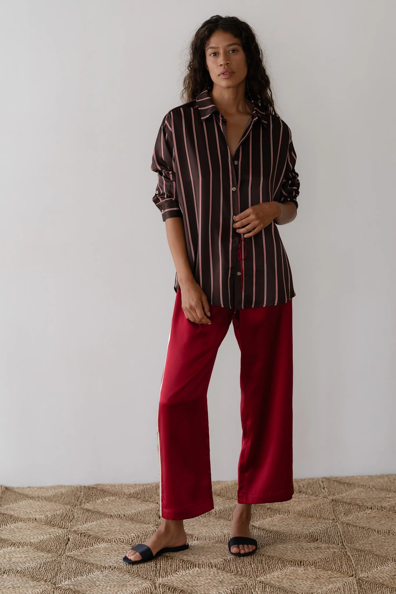 A person with long hair stands against a white wall, wearing a striped, long-sleeve shirt and striking red "The Silk Stripe Crop Pant" by Donni. They are on a woven rug and wear black slip-on shoes, embodying the effortless style of Los Angeles in their 100% silk ensemble.