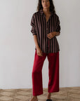 A person with long hair stands against a white wall, wearing a striped, long-sleeve shirt and striking red "The Silk Stripe Crop Pant" by Donni. They are on a woven rug and wear black slip-on shoes, embodying the effortless style of Los Angeles in their 100% silk ensemble.