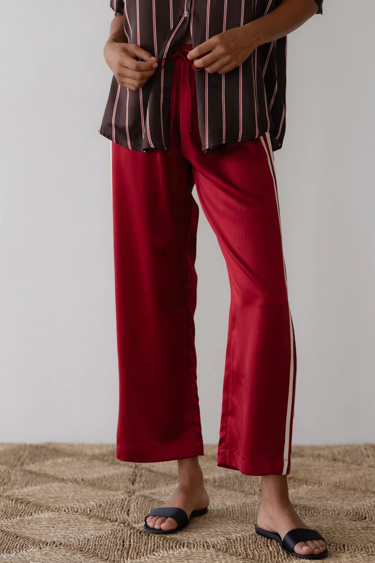 In Los Angeles, someone stands on a textured beige carpet, wearing red satin pants made of 100% silk with white side stripes from Donni's The Silk Stripe Crop Pant collection, paired with a brown and white striped shirt. Their look is finished with black slip-on sandals.