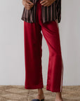 In Los Angeles, someone stands on a textured beige carpet, wearing red satin pants made of 100% silk with white side stripes from Donni's The Silk Stripe Crop Pant collection, paired with a brown and white striped shirt. Their look is finished with black slip-on sandals.