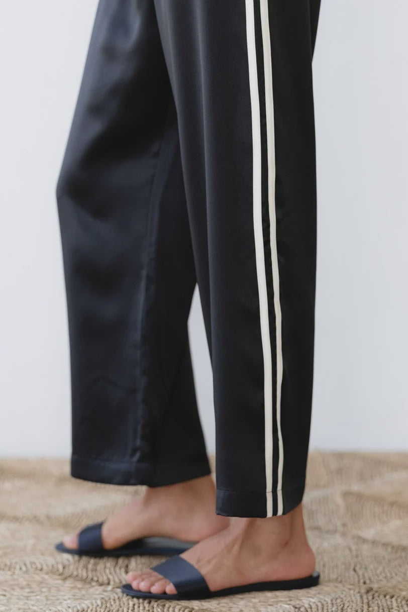 A person wearing The Silk Stripe Crop Pant by Donni in black with white silk side stripes and dark blue slide sandals stands on a woven rug.