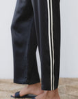 A person wearing The Silk Stripe Crop Pant by Donni in black with white silk side stripes and dark blue slide sandals stands on a woven rug.