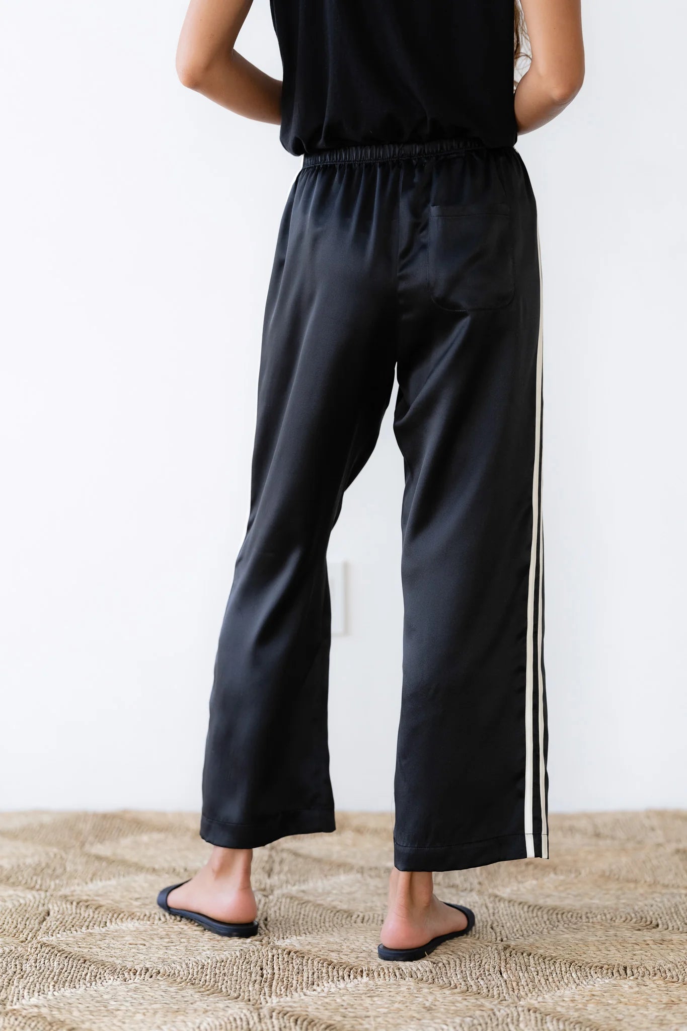 Dressed in the elegant Silk Stripe Crop Pant by Donni, a person stands on a woven rug, showcasing style with the 100% silk satin pants. The pants feature a chic single white stripe down each side, perfectly capturing effortless Los Angeles-inspired fashion as they stand with their arms at their sides, facing away from the camera.