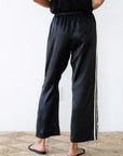 Dressed in the elegant Silk Stripe Crop Pant by Donni, a person stands on a woven rug, showcasing style with the 100% silk satin pants. The pants feature a chic single white stripe down each side, perfectly capturing effortless Los Angeles-inspired fashion as they stand with their arms at their sides, facing away from the camera.