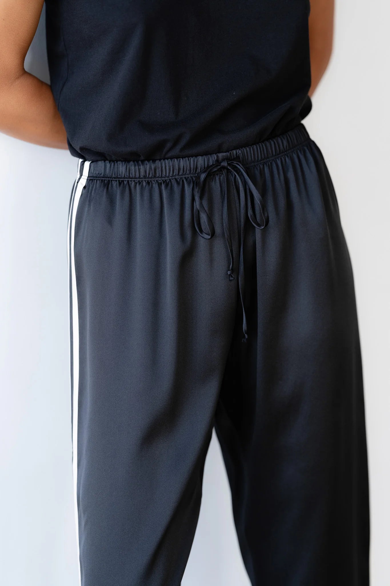 A person stands wearing Donni&#39;s The Silk Stripe Crop Pant, channeling a relaxed Los Angeles vibe with its sleek dark fabric and elegant white silk side stripes. The adjustable drawstring waist and comfortable fit are an ideal match with a black t-shirt.