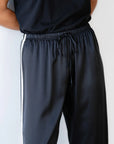 A person stands wearing Donni's The Silk Stripe Crop Pant, channeling a relaxed Los Angeles vibe with its sleek dark fabric and elegant white silk side stripes. The adjustable drawstring waist and comfortable fit are an ideal match with a black t-shirt.