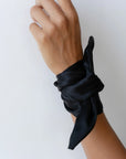 A hand against a white background is adorned with an elegant accessory from Donni: The Silk Bow, crafted from 100% black silk and tied gracefully around the wrist.