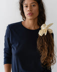 A person with long hair tied in a low ponytail with Donni's "The Silk Bow" in cream is wearing a navy blue long-sleeve shirt and beige pants. They are standing against a plain white background, gazing directly at the camera.