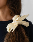 The image captures a close-up of a person with long, wavy brown hair styled in a low ponytail, adorned with The Silk Bow by Donni, featuring an elegant, 100% silk design in cream. This accessory beautifully complements the dark navy top, highlighting both the hairstyle and The Silk Bow.