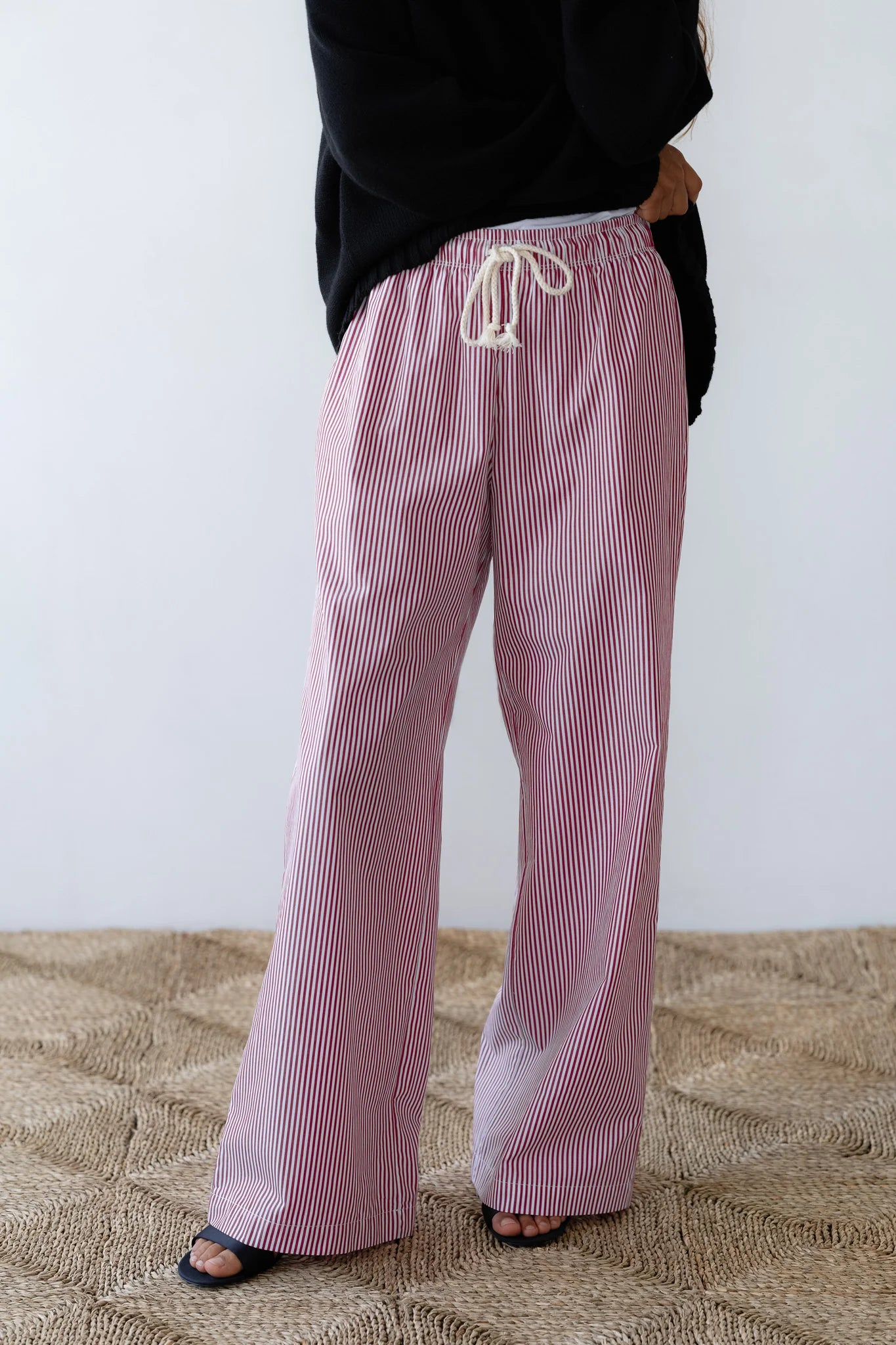 A person wears The Stripe Pop Pant by Donni, featuring wide-legged, red and white striped cotton poplin with a drawstring waist. Paired with a black top and open-toe sandals on a textured floor against a neutral wall, their upper face is obscured.