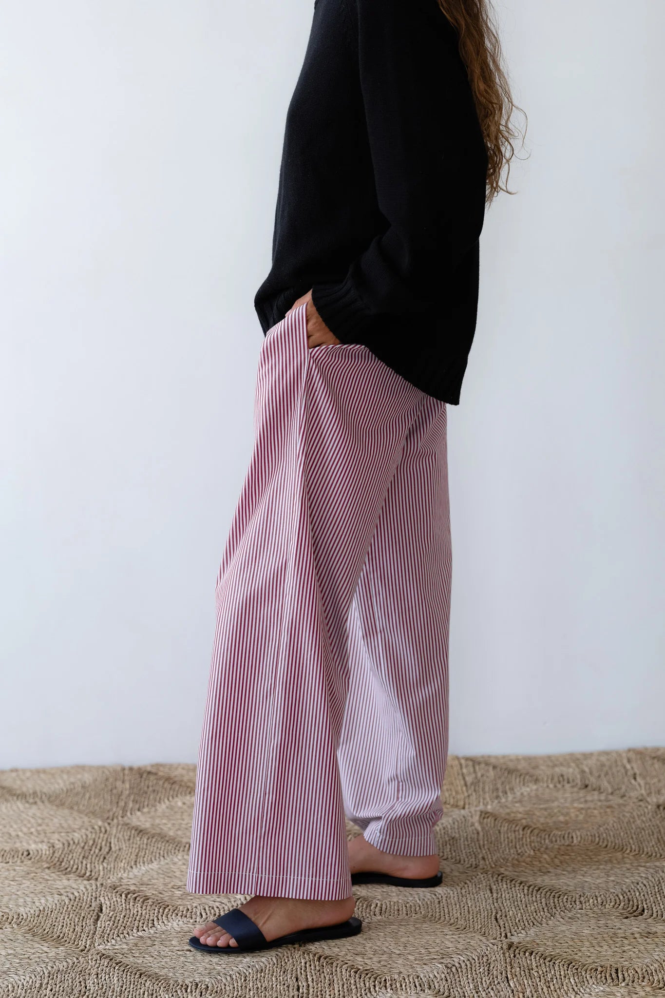 A person stands on a woven rug wearing Donni's The Stripe Pop Pant in red-and-white stripes and a black long-sleeve top, with only the lower half visible, completed by black slide sandals.