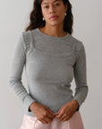 A person with long hair wears a slim-fit, light gray "The Baby Rib Long Sleeve" by Donni and a pink skirt, standing against a plain background, looking forward with hands clasped in front.