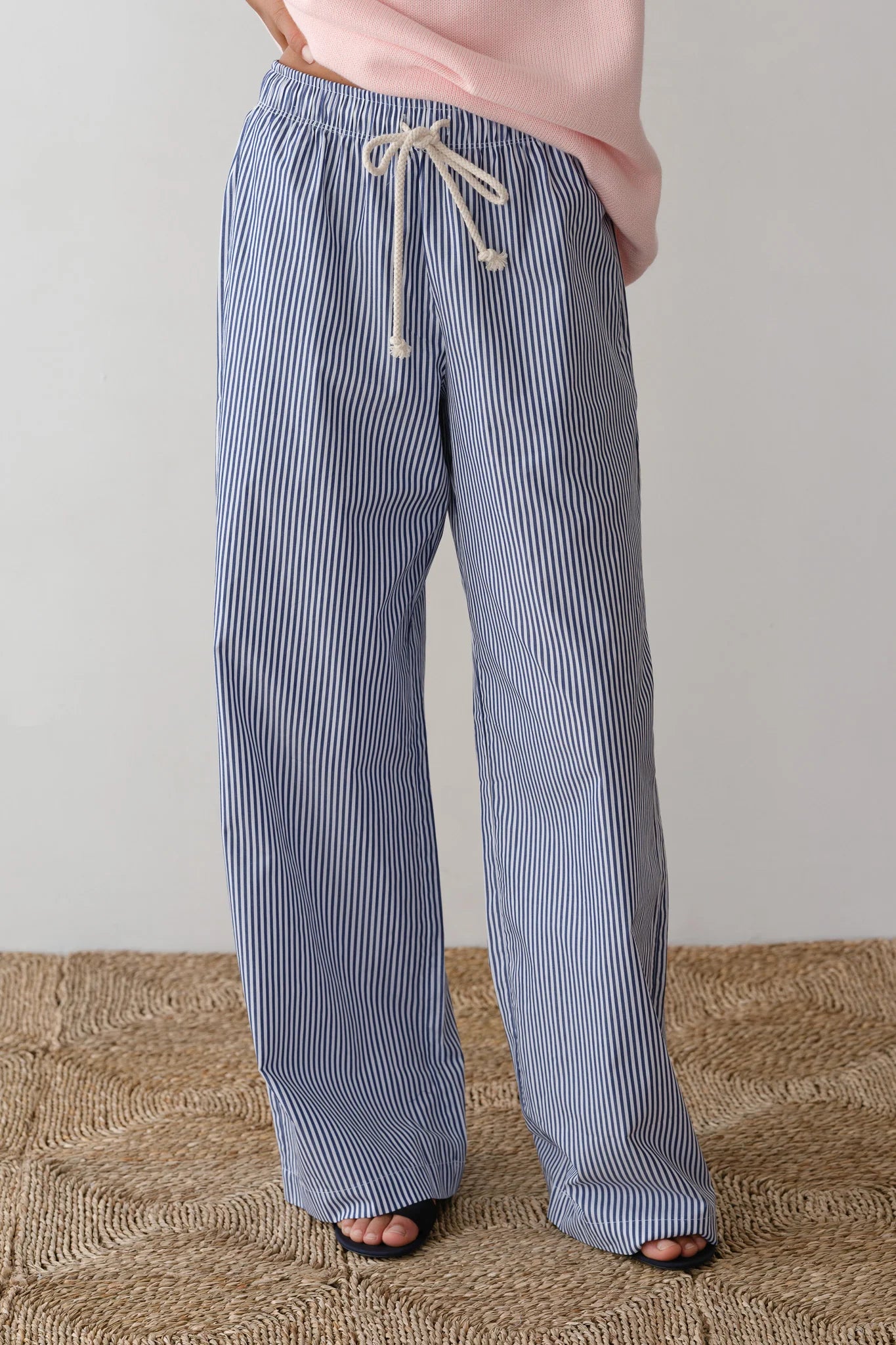 A person stands gracefully on a textured beige rug wearing Donni's The Stripe Pop Pant, with its stylish wide-leg fit and drawcord waist. Their look is complemented by open-toe sandals, and the top of their pink shirt peeks out.