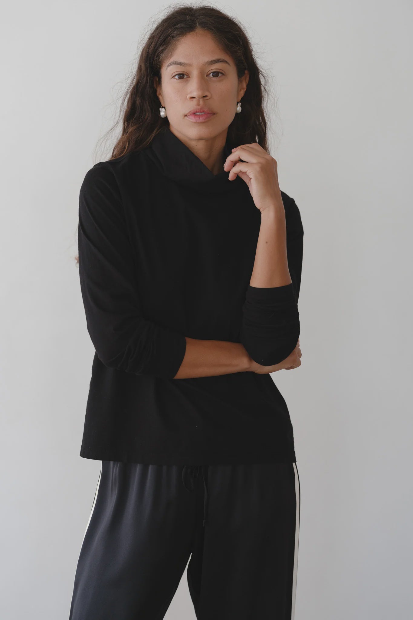 A woman with long curly hair wears The Jersey Turtleneck by Donni, paired with matching black pants featuring white side stripes. Made in Los Angeles, she stands against a plain light background, one arm crossed and the other touching her chin, exuding a calm and confident expression.