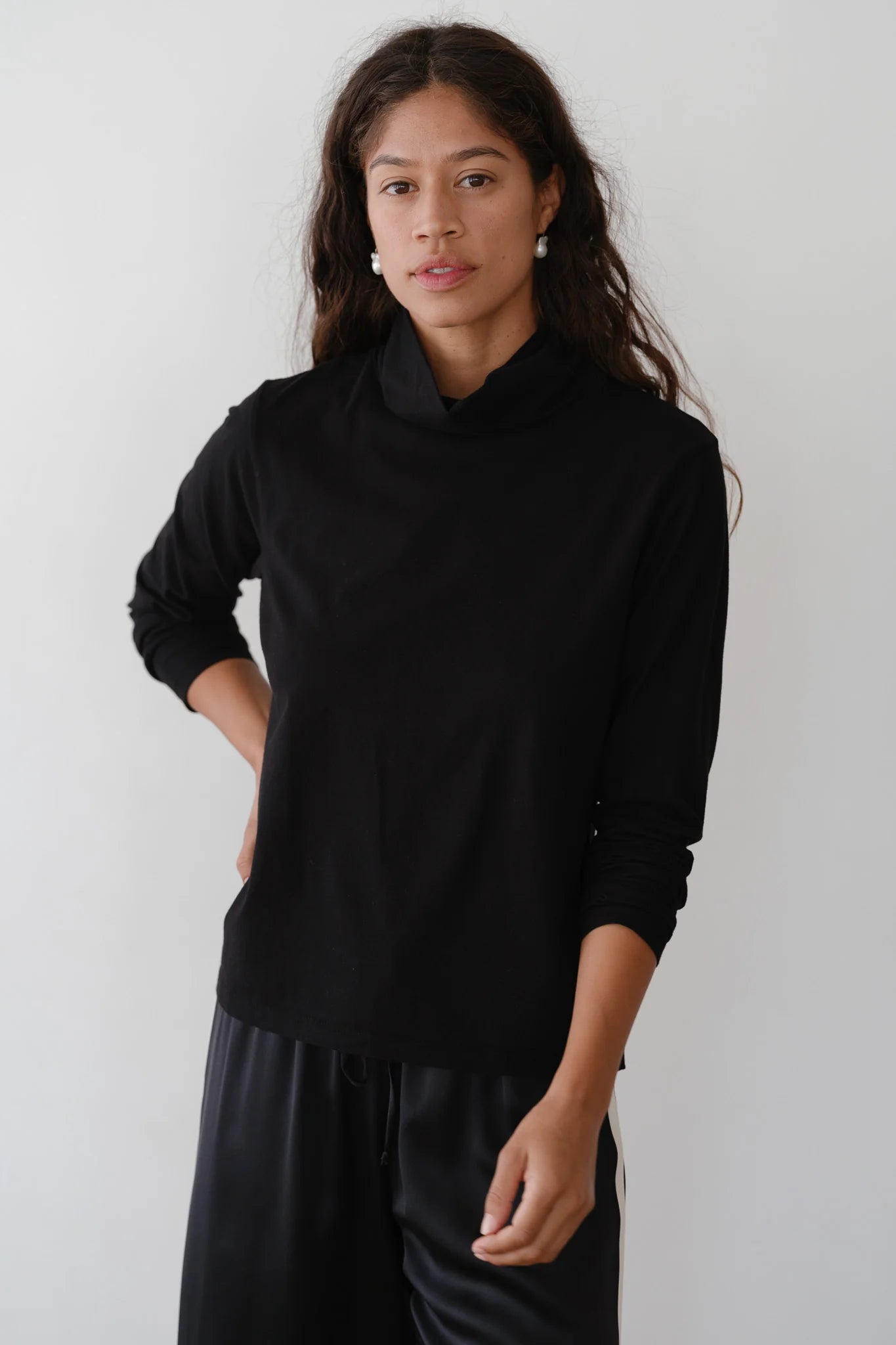 A person with long hair stands against a plain white background, wearing The Jersey Turtleneck and matching pants by Donni. Their left hand rests on their hip, and they have a neutral expression. The outfit's relaxed fit adds to the overall sleek look.