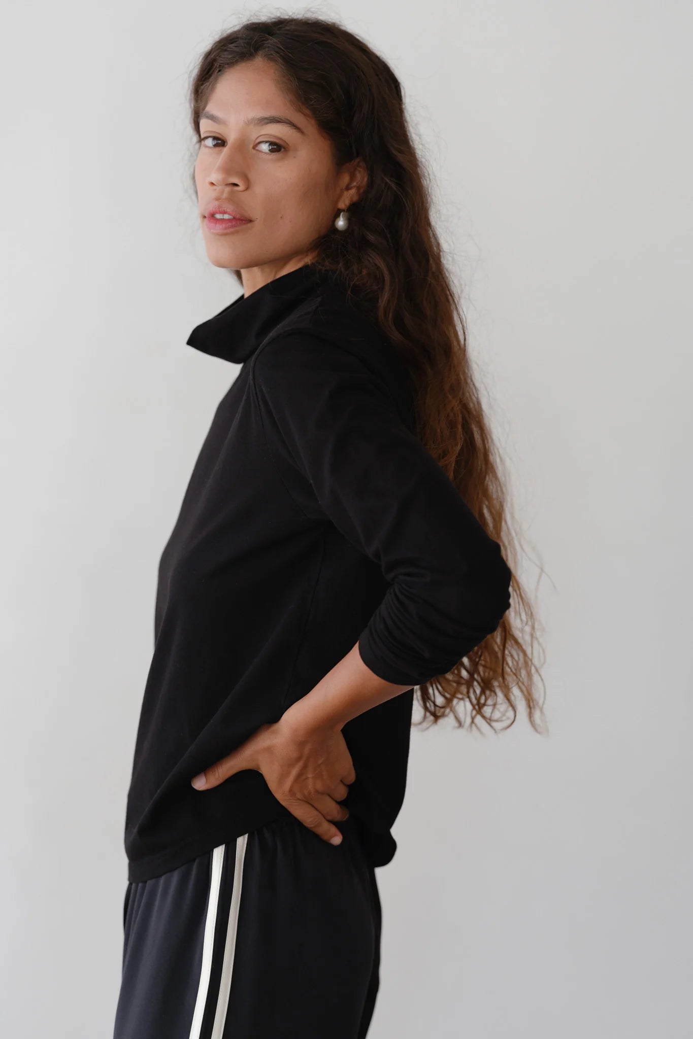 A person with long hair stands sideways, wearing The Jersey Turtleneck by Donni and black pants featuring white stripes. They have one hand on their hip and are looking at the camera against a plain background.