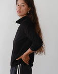 A person with long hair stands sideways, wearing The Jersey Turtleneck by Donni and black pants featuring white stripes. They have one hand on their hip and are looking at the camera against a plain background.