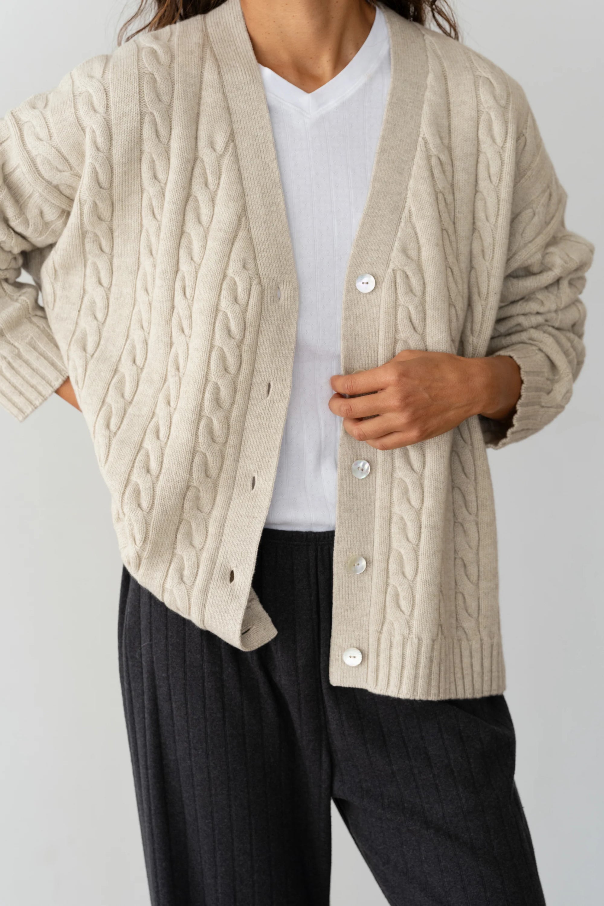 Someone in a cream-colored Donni The Cable Vneck Cardigan over a white shirt and dark gray pants, with the cardigan open to show part of the shirt, rests one hand on their stomach.