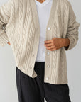 Someone in a cream-colored Donni The Cable Vneck Cardigan over a white shirt and dark gray pants, with the cardigan open to show part of the shirt, rests one hand on their stomach.