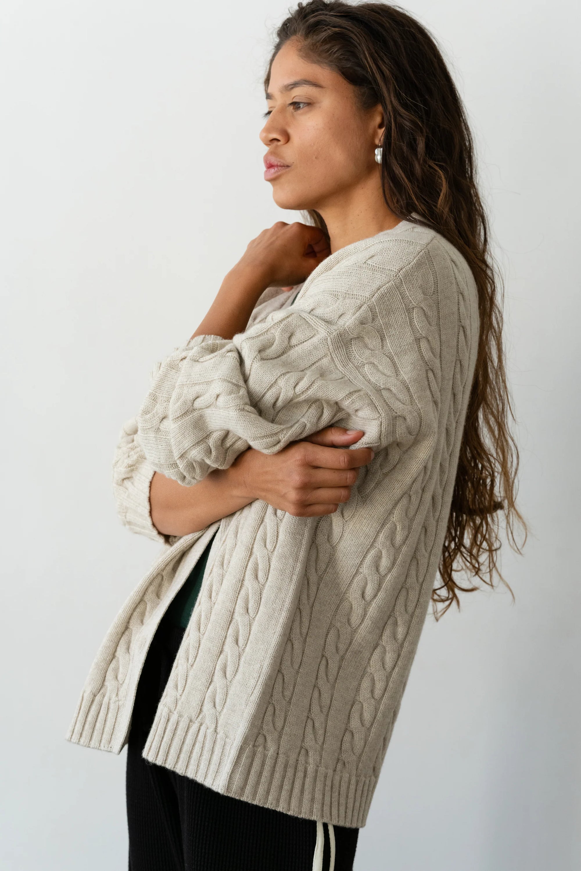 A person with long, wavy hair stands in profile, wearing The Cable Vneck Card by Donni in beige. With their arms crossed, they gaze thoughtfully into the distance against a plain, light backdrop.