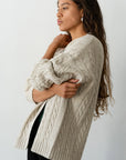 A person with long, wavy hair stands in profile, wearing The Cable Vneck Card by Donni in beige. With their arms crossed, they gaze thoughtfully into the distance against a plain, light backdrop.